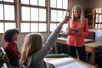 Chica caucásica levantando la mano mientras está sentada en su escritorio durante la lección con la maestra caucásica. Educación primaria distanciamiento social seguridad sanitaria durante la pandemia del Coronavirus Covid19. - foto de stock