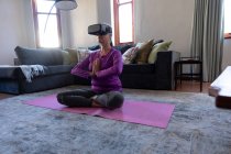 Kaukasische Frau genießt Zeit zu Hause, soziale Distanzierung und Selbstisolierung in Quarantäne, übt im Wohnzimmer, trägt VR-Headset, sitzt im Schneidersitz auf dem Boden, meditiert. — Stockfoto