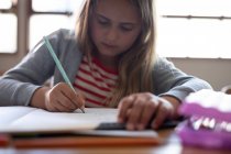 Menina branca escrevendo em um livro enquanto sentada em sua mesa na escola. Educação primária distanciamento social segurança sanitária durante Covid19 pandemia de coronavírus. — Fotografia de Stock