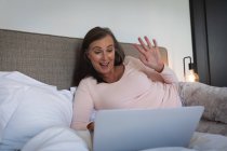Femme caucasienne appréciant le temps à la maison, la distance sociale et l'isolement personnel en quarantaine verrouillée, couchée sur le lit dans la chambre, à l'aide d'un ordinateur portable, saluant lors d'un appel vidéo. — Photo de stock