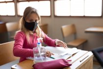 Chica caucásica usando mascarilla mientras está sentada en el escritorio y desinfectando sus manos. Educación primaria distanciamiento social seguridad sanitaria durante la pandemia del Coronavirus Covid19. - foto de stock