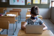Garoto caucasiano usando uma máscara facial, usando laptop enquanto estava sentado em sua mesa na escola. Educação primária distanciamento social segurança sanitária durante Covid19 pandemia de coronavírus. — Fotografia de Stock