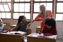 Eine Gruppe multiethnischer Kinder sitzt mit einer kaukasischen Lehrerin auf ihrem Schreibtisch im Klassenzimmer der Schule. Grundschulbildung soziale Distanzierung der Gesundheitssicherheit während der Covid19 Coronavirus-Pandemie. — Stockfoto