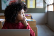 Thoughtful mixed race boy using laptop while sitting on his desk in class at school. Primary education social distancing health safety during Covid19 Coronavirus pandemic. — Stock Photo