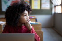 Garçon métis réfléchi utilisant un ordinateur portable alors qu'il était assis sur son bureau en classe à l'école. Enseignement primaire distanciation sociale sécurité sanitaire pendant la pandémie de coronavirus Covid19. — Photo de stock