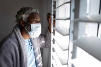 Afroamerikanische Senioren, die in einem Schlafzimmer stehen, eine Gesichtsmaske tragen, durch ein Fenster schauen, soziale Distanzierung und Selbstisolierung in Quarantäne — Stockfoto