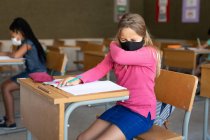 Caucasian girl sitting at desk wearing face mask in classroom, covering her face while sneezing. Primary education social distancing health safety during Covid19 Coronavirus pandemic. — Stock Photo
