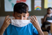 Gemischter Junge mit Gesichtsmaske, während er in der Schule auf seinem Schreibtisch sitzt. Grundschulbildung soziale Distanzierung der Gesundheitssicherheit während der Covid19 Coronavirus-Pandemie. — Stockfoto