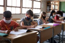 Multi ethnic group of elementary school children sitting at desks wearing face masks in classroom. Primary education social distancing health safety during Covid19 Coronavirus pandemic. — Stock Photo