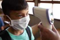 Mixed race boy wearing face mask getting his temperature measured in an elementary school. Primary education social distancing health safety during Covid19 Coronavirus pandemic. — Stock Photo