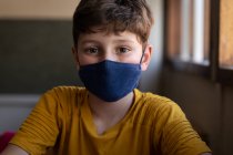 Portrait d'un garçon caucasien assis au bureau portant un masque facial en classe. Enseignement primaire distanciation sociale sécurité sanitaire pendant la pandémie de coronavirus Covid19. — Photo de stock