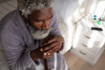 Афроамериканський старший чоловік сидить на ліжку в спальні, спочиваючи голову на тростині, соціальній відстані і самоізоляції в карантинному блокуванні. — стокове фото