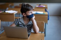 Menino caucasiano cuidadoso usando uma máscara facial, usando laptop enquanto sentado em sua mesa na sala de aula na escola. Educação primária distanciamento social segurança sanitária durante Covid19 pandemia de coronavírus. — Fotografia de Stock
