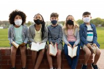 Group of multi ethnic kids wearing face masks reading books while sitting on the wall during a break. Primary education social distancing health safety during Covid19 Coronavirus pandemic. — Stock Photo