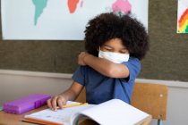 Gemischter Junge mit Gesichtsmaske sitzt im Klassenzimmer am Schreibtisch und verdeckt sein Gesicht, während er niest. Grundschulbildung soziale Distanzierung der Gesundheitssicherheit während der Covid19 Coronavirus-Pandemie. — Stockfoto