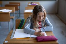 Белая девушка пишет в книге, сидя на столе в школе. Начальное образование Социальное дистанцирование безопасности здоровья во время пандемии Coronavirus Covid19. — стоковое фото