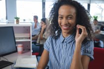 Portrait of smart mixed race businesswoman sitting at desk wearing phone headset smiling to camera, with colleagues in the background. Creative business professionals working in busy modern office. — Stock Photo