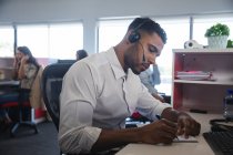 Smarte, lässig gekleidete gemischte Geschäftsleute sitzen mit Kopfhörern am Schreibtisch und schreiben Notizen, im Hintergrund Kollegen. Kreative Geschäftsprofis arbeiten in geschäftigen modernen Büros. — Stockfoto