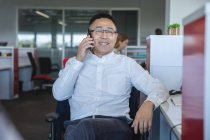 Inteligente casualmente vestido asiático negocio masculino creativo usando gafas sentadas en un escritorio sonriendo, hablando en el teléfono inteligente. Profesional creativo de negocios que trabaja en una oficina moderna. - foto de stock