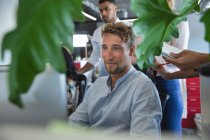 Smart casually dressed Caucasian male business creative sitting at a desk smiling, with colleagues gathered around him taking notes. Creative business professionals working in a busy modern office. — Stock Photo