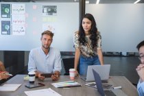 Asian businesswoman standing to address colleagues during a team meeting, a Caucasian male colleague sitting beside her listening. Creative business professionals working in a busy modern office. — Stock Photo
