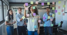 Multi-ethnic male and female business team brainstorming together, a mixed race woman writing on sticky notes stuck to a glass wall. Creative business professionals working in a busy modern office. — Stock Photo