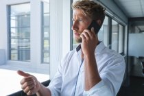 Smart casually dressed Caucasian male business creative with blonde hair and beard looking out of window, talking on smartphone. Creative business professional working in a modern office. — Stock Photo