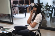 Frau mit gemischter Rasse, die in einem lässigen Büro arbeitet, ihr Headset trägt und auf den virtuellen Bildschirm blickt. Soziale Distanzierung am Arbeitsplatz während der Coronavirus Covid 19 Pandemie. — Stockfoto
