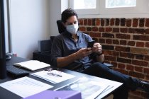 Kaukasischer Mann, der in einem lässigen Büro arbeitet, sein Smartphone benutzt und Gesichtsmaske trägt. Soziale Distanzierung am Arbeitsplatz während der Coronavirus Covid 19 Pandemie. — Stockfoto