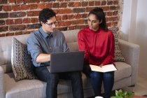 Mixed race woman and Caucasian man working in a casual office, sitting on a sofa, using a laptop computer and talking. Creative business professionals working in a busy modern office. — Stock Photo