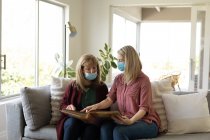 Senior Caucasian woman spending time at home with her adult daughter, sitting on couch, wearing face masks and reading a book. Social distancing during Covid 19 Coronavirus quarantine. — Stock Photo