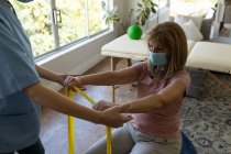 Femme caucasienne âgée à la maison visitée par une infirmière caucasienne, étirant les bras à l'aide d'une bande d'exercice, portant un masque facial. Soins médicaux à domicile pendant la quarantaine du coronavirus Covid 19. — Photo de stock