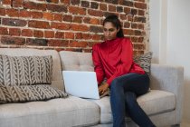 Mujer de raza mixta trabajando en una oficina informal, sentada en un sofá, usando una computadora portátil. Distanciamiento social en el lugar de trabajo durante la pandemia de Coronavirus Covid 19. - foto de stock