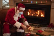 Homme caucasien âgé à la maison habillé comme le Père Noël, portant un masque, assis sur une chaise près de la cheminée, donnant des cadeaux. Distance sociale pendant le confinement en quarantaine du coronavirus Covid 19. — Photo de stock