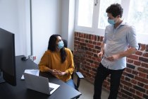 Mixed race woman and Caucasian man working in a casual office, wearing face masks and talking. Social distancing in the workplace during Coronavirus Covid 19 pandemic. — Stock Photo