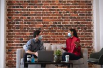 Mixed race woman and Caucasian man working in a casual office, wearing face masks, sitting on a sofa and talking. Social distancing in the workplace during Coronavirus Covid 19 pandemic. — Stock Photo