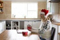 Mujer caucásica pasar tiempo en casa, sentado en la cocina en Navidad con sombrero de Santa, el uso de ordenador portátil con regalos en la mesa. Distanciamiento social durante la cuarentena del Coronavirus Covid 19. - foto de stock