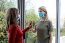 Senior Caucasian woman and her adult daughter at home, wearing face masks and greeting each other by touching hands. Social distancing, health and hygiene during Covid 19 Coronavirus pandemic. — Stock Photo