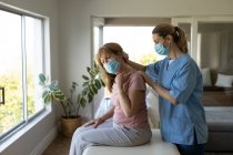 Senior Caucasian woman at home visited by Caucasian female nurse, stretching her neck, wearing face masks. Medical care at home during Covid 19 Coronavirus quarantine. — Stock Photo
