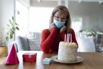 Mulher caucasiana sênior passando tempo em casa, sentada em sua sala de estar com um bolo de aniversário, usando máscara facial. Distanciamento social durante o bloqueio de quarentena do Covid 19 Coronavirus. — Fotografia de Stock