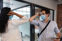 Змішана расова жінка і кавказький чоловік, які працюють у повсякденному офісі, носять маску обличчя і торкаються один одного ліктями. Суспільна дистанція на робочому місці під час Коронавірусу Ковід 19 пандемії. — стокове фото