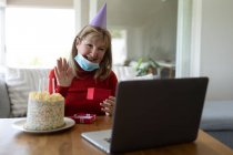 Mujer mayor caucásica pasar tiempo en casa, sentada en su sala de estar con un pastel de cumpleaños, con máscara facial y el uso de un ordenador portátil. Distanciamiento social durante el bloqueo de cuarentena del Coronavirus Covid 19. - foto de stock