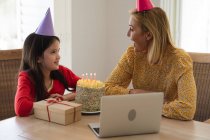 Mujer caucásica y su hija pasan tiempo en casa juntos, celebrando cumpleaños, usando una computadora portátil, haciendo una videollamada. Distanciamiento social durante el bloqueo de cuarentena del Coronavirus Covid 19. - foto de stock