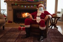 Senior Caucasian man at home dressed as Father Christmas, wearing face mask, sitting on a chair by fireplace. Social distancing during Covid 19 Coronavirus quarantine lockdown. — Stock Photo