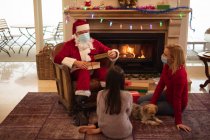 Старший белый мужчина, его взрослая дочь и внучка дома, одетые как Дед Мороз, в маске для лица, дарят подарки. Социальное дистанцирование во время изоляции коронавируса Covid 19. — стоковое фото