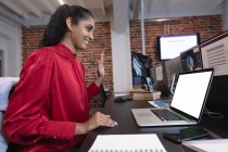 Mujer de raza mixta trabajando en una oficina informal, sentada en el escritorio, usando una computadora portátil, saludando. Distanciamiento social en el lugar de trabajo durante la pandemia de Coronavirus Covid 19. - foto de stock