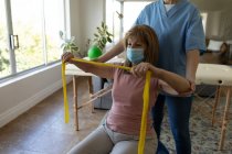 Senior Caucasian woman at home visited by Caucasian female nurse, stretching arms using exercise band, wearing face mask. Medical care at home during Covid 19 Coronavirus quarantine. — Stock Photo