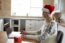 Femme caucasienne passant du temps à la maison, assis dans la cuisine à Noël portant le chapeau de Père Noël, en utilisant un ordinateur portable avec des cadeaux sur la table. Distance sociale pendant la quarantaine du coronavirus Covid 19. — Photo de stock
