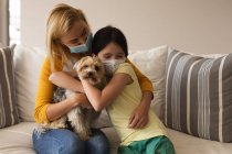 Mulher caucasiana e sua filha passam o tempo em casa juntos, usando máscaras, abraçando seu cão. Distanciamento social durante o bloqueio de quarentena do Covid 19 Coronavirus. — Fotografia de Stock