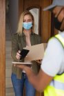 Caucasian woman spending time at home, wearing face mask, receiving a package from delivery man and paying by smartphone. Social distancing during Covid 19 Coronavirus quarantine lockdown. — Stock Photo